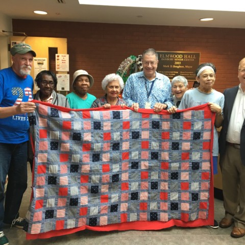 Mayor Mark posing with seniors at Elmwood Hall showing off one of the quilts on sale for the Craft and Bake Sale